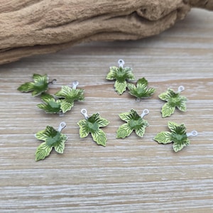 May include: A collection of ten green metal leaf charms with silver jump rings. The leaves have a detailed design and a glossy finish.