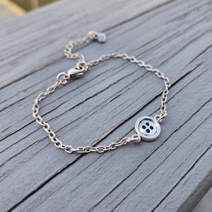 Puede incluir: Un brazalete de cadena de plata con un dije de botón de plata. El botón tiene cuatro agujeros.