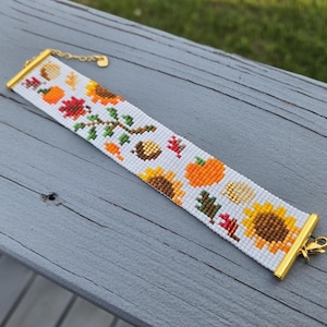 Peut inclure: Bracelet de perles blanches avec un motif de r&eacute;colte d'automne. Le bracelet a un fermoir et une cha&icirc;ne dor&eacute;s. Le motif comprend des tournesols, des citrouilles, des feuilles et des glands.