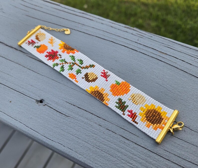 Peut inclure: Bracelet de perles blanches avec un motif de r&eacute;colte d'automne. Le bracelet a un fermoir et une cha&icirc;ne dor&eacute;s. Le motif comprend des tournesols, des citrouilles, des feuilles et des glands.