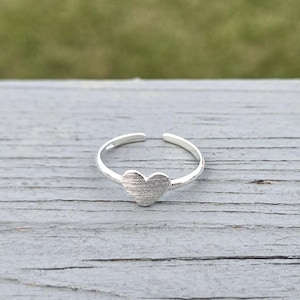 May include: A silver toe ring with a heart-shaped design. The heart has a textured surface.