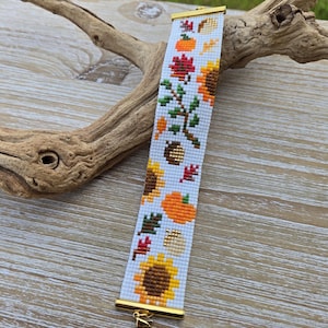 Peut inclure: Un bracelet de perles sur le th&egrave;me de l'automne, avec des tournesols, des citrouilles et des feuilles orange, jaunes, rouges et vertes sur fond blanc. Le bracelet a des extr&eacute;mit&eacute;s en m&eacute;tal dor&eacute; et un fermoir.