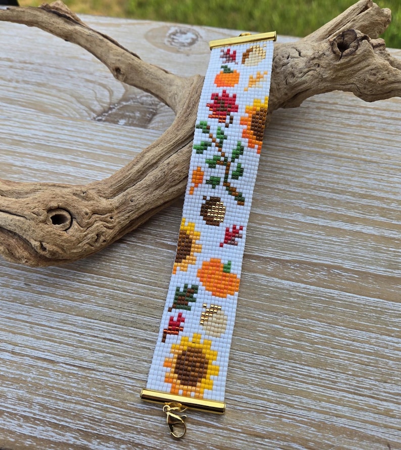Peut inclure: Un bracelet de perles sur le th&egrave;me de l'automne, avec des tournesols, des citrouilles et des feuilles orange, jaunes, rouges et vertes sur fond blanc. Le bracelet a des extr&eacute;mit&eacute;s en m&eacute;tal dor&eacute; et un fermoir.