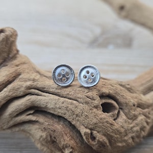 May include: A pair of small, circular silver-toned stud earrings shaped like classic four-hole sewing buttons. The earrings feature a textured, matte metallic finish with a slightly raised rim and four distinct circular indentations in the center.