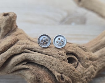 May include: A pair of small, circular silver-toned stud earrings shaped like classic four-hole sewing buttons. The earrings feature a textured, matte metallic finish with a slightly raised rim and four distinct circular indentations in the center.