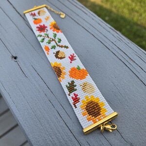 Peut inclure: Bracelet blanc perl&eacute; avec des accents dor&eacute;s et un fermoir. Le bracelet pr&eacute;sente un motif r&eacute;p&eacute;t&eacute; d'art pixel sur le th&egrave;me de l'automne, comprenant des tournesols, des citrouilles et des feuilles. Le bracelet mesure environ 18 cm de long.