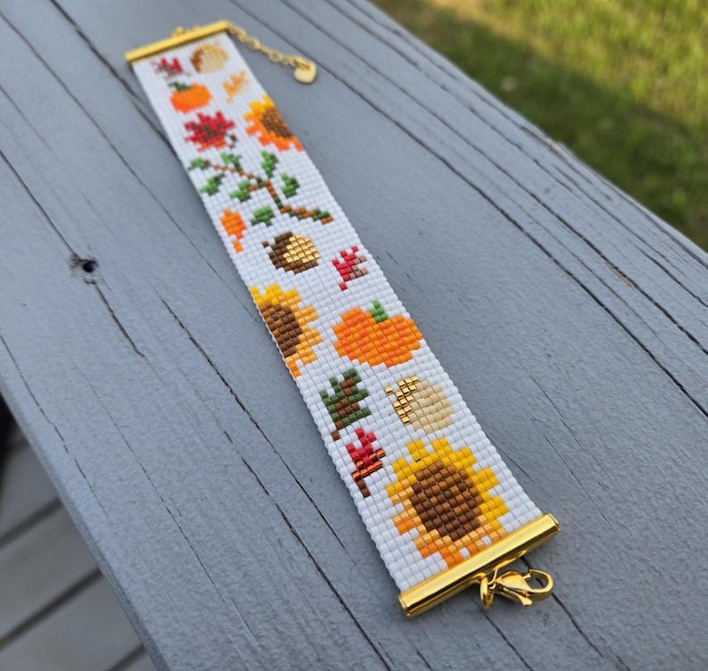 Peut inclure: Bracelet blanc perl&eacute; avec des accents dor&eacute;s et un fermoir. Le bracelet pr&eacute;sente un motif r&eacute;p&eacute;t&eacute; d'art pixel sur le th&egrave;me de l'automne, comprenant des tournesols, des citrouilles et des feuilles. Le bracelet mesure environ 18 cm de long.