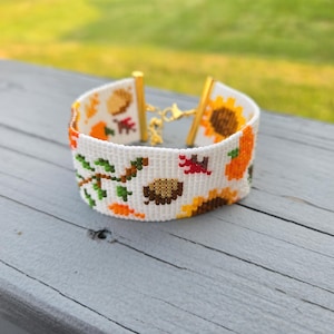 Peut inclure: Bracelet blanc orn&eacute; d'un motif de r&eacute;colte d'automne. Le bracelet est dot&eacute; d'un fermoir dor&eacute; et est d&eacute;cor&eacute; de tournesols, de citrouilles, de feuilles et de glands en orange, jaune, marron et vert.