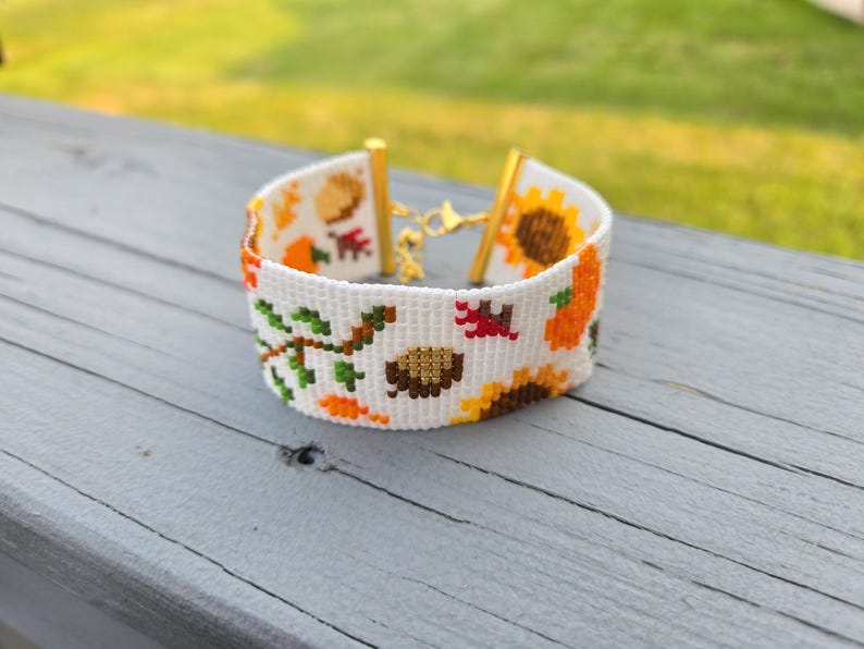 Peut inclure: Bracelet blanc orn&eacute; d'un motif de r&eacute;colte d'automne. Le bracelet est dot&eacute; d'un fermoir dor&eacute; et est d&eacute;cor&eacute; de tournesols, de citrouilles, de feuilles et de glands en orange, jaune, marron et vert.