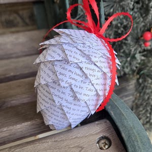 May include: A white paper ornament shaped like a pine cone with a red ribbon. The ornament is covered in handwritten text, including the words "take my hand" and "in love".