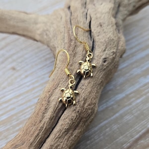 May include: A pair of gold-toned drop earrings featuring small, detailed turtle charms. Each earring is suspended from a simple gold-toned hook wire, showcasing a polished finish and a clear, segmented shell pattern on the back of each turtle.
