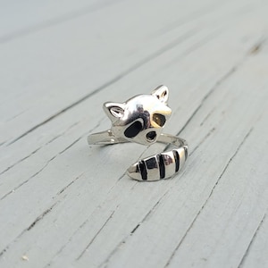 May include: A silver ring with a raccoon design. The raccoon's face is on the front of the ring, and its tail wraps around the band.