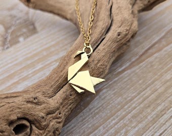 May include: A gold-colored origami crane pendant necklace. The pendant is suspended from a delicate gold chain. The necklace is displayed on a piece of driftwood against a light wood background.