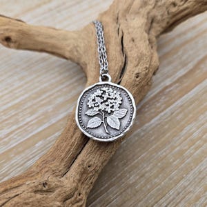 May include: Silver pendant necklace with a floral design. The round pendant features a raised image of a flower and leaves, encircled by a dotted border. The necklace hangs from a delicate chain. The background is natural wood.