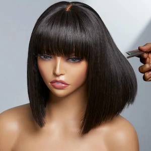 May include: A mannequin head with a black, blunt-cut bob wig. The wig features a straight fringe and a shoulder-length cut. A hand holds a comb near the hair, suggesting styling or cutting. The mannequin has realistic features and a neutral expression.