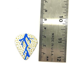May include: A guitar pick with a white background, a blue lightning bolt design, and yellow dots. A metal ruler is next to the pick, showing measurements in inches and millimeters.