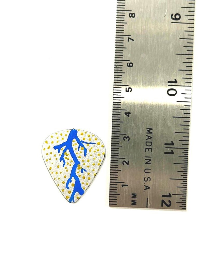 May include: A guitar pick with a white background, a blue lightning bolt design, and yellow dots. A metal ruler is next to the pick, showing measurements in inches and millimeters.