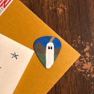 Puede incluir: Una púa de guitarra con una escena pintada a mano de un fantasma blanco de pie en un campo verde con montañas marrones al fondo bajo un cielo azul. Un pequeño arcoíris se arquea sobre la cabeza del fantasma, que tiene dos ojos negros.