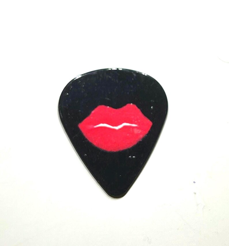 May include: Black guitar plectrum with a red lip design. The glossy plectrum has a teardrop shape and a white background. The red lips are centered on the pick.