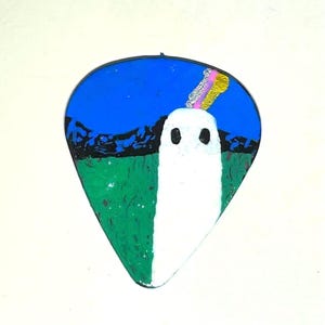 Może przedstawiać: Kostka do gitary z namalowanym wzorem. Kostka przedstawia białą postać ducha na zielono-niebieskim tle. Tęczowy łuk rozciąga się z głowy ducha. Kostka ma kształt łzy.