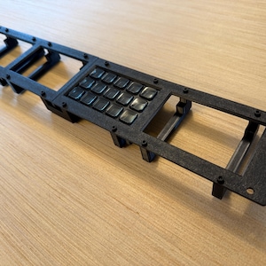 May include: Black, rectangular keyboard frame with a grid of 16 square keys. The frame has a textured surface and is secured with small screws. The design suggests a custom or specialized keyboard component.