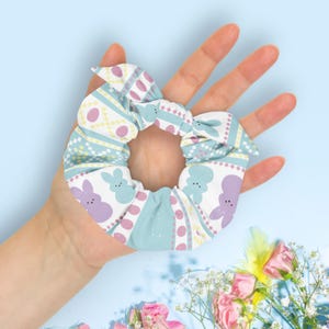 May include: A pastel-colored scrunchie with an Easter theme. The fabric features a pattern of purple and blue bunnies, pink eggs, and geometric shapes on a light blue background. The scrunchie is held in a hand.