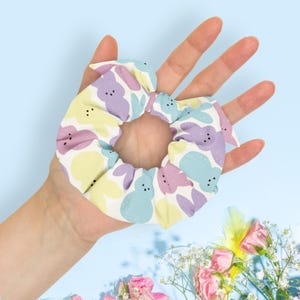 May include: A pastel-colored scrunchie featuring a repeating pattern of Easter bunny shapes in light blue, lavender, and yellow on a white background. The scrunchie is held in a hand against a light blue backdrop, with flowers in the lower right corner.