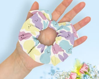 May include: A pastel-coloured scrunchie with a repeating pattern of Easter bunny shapes in light blue, lavender, and yellow on a white background. The scrunchie is held in a hand against a light blue backdrop, with flowers in the lower right corner.