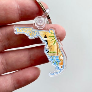 May include: A clear acrylic keychain shaped like the state of Florida with a colorful illustration of a lighthouse, palm trees, and oranges. The keychain has a silver metal ring and a clear plastic swivel.
