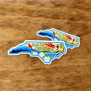 May include: Two North Carolina state outline stickers with a blue, white, and yellow design. The stickers feature a lighthouse, a red cardinal, a white flower, and a red airplane. The text "North Carolina" is written in a stylized font.