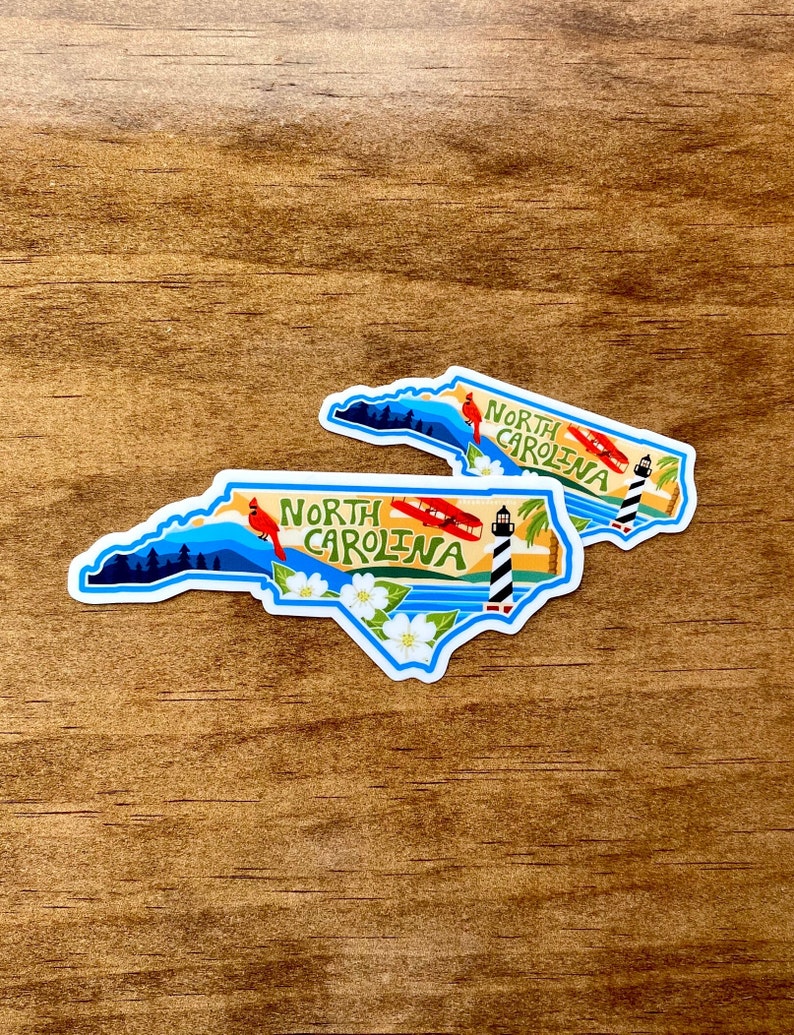 May include: Two North Carolina state outline stickers with a blue, white, and yellow design. The stickers feature a lighthouse, a red cardinal, a white flower, and a red airplane. The text "North Carolina" is written in a stylized font.