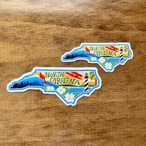 May include: Two North Carolina state outline stickers with a blue outline and a white interior. The stickers feature a red cardinal, a lighthouse, a white flower, and the text "North Carolina".