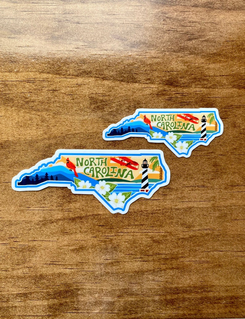 May include: Two North Carolina state outline stickers with a blue outline and a white interior. The stickers feature a red cardinal, a lighthouse, a white flower, and the text "North Carolina".
