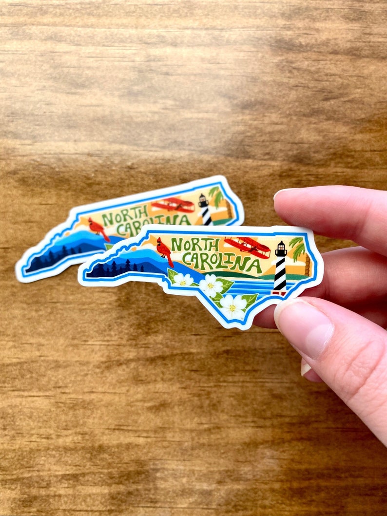May include: Two North Carolina state outline stickers with a blue border. The stickers feature a cardinal, a lighthouse, a plane, and white flowers. The text "North Carolina" is written on each sticker.