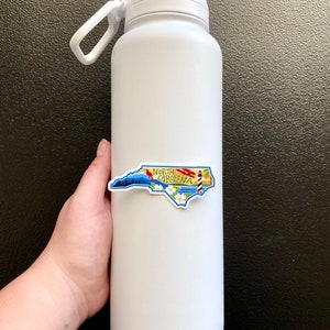 May include: White insulated water bottle with a North Carolina state outline sticker. The sticker features a lighthouse, a cardinal, and the words "North Carolina".