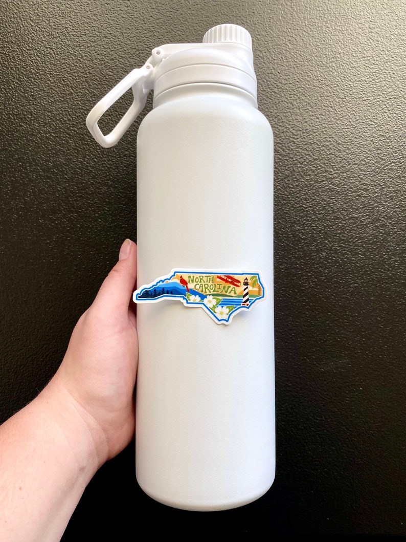 May include: White insulated water bottle with a North Carolina state outline sticker. The sticker features a lighthouse, a cardinal, and the words "North Carolina".