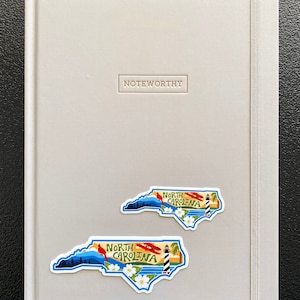 May include: A gray notebook with the word "Noteworthy" embossed on the front cover. Two North Carolina state outline stickers are on the front cover. The stickers feature a blue sky, mountains, a lighthouse, a cardinal, and white flowers. The text "North Carolina" is printed on each sticker.