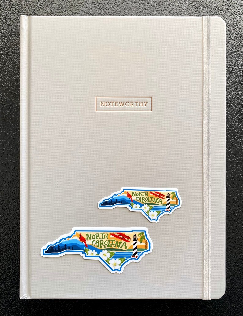 May include: A gray notebook with the word "Noteworthy" embossed on the front cover. Two North Carolina state outline stickers are on the front cover. The stickers feature a blue sky, mountains, a lighthouse, a cardinal, and white flowers. The text "North Carolina" is printed on each sticker.