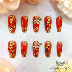 May include: Ten red and gold press-on nails with rhinestones and gold accents. The nails are long and have a coffin shape.