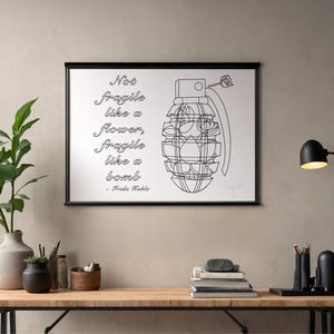May include: Black framed art print featuring a line drawing of a grenade with the quote "Not fragile like a flower, fragile like a bomb" by Frida Kahlo. The art is displayed above a wooden desk with plants and a lamp.