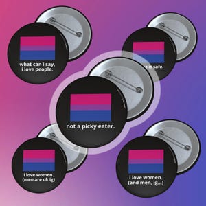 May include: A collection of black round buttons with a pin back. Each button features a bisexual pride flag design in pink, purple, and blue. Some buttons have text: "what can I say, I love people," "not a picky eater," and "I love women."
