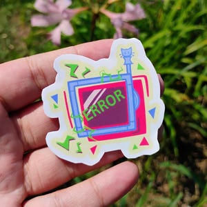May include: A die-cut sticker featuring a retro television set with a pink screen displaying the word ERROR in green text. The design includes a small cat silhouette on top of the antenna, surrounded by abstract green and blue geometric shapes.