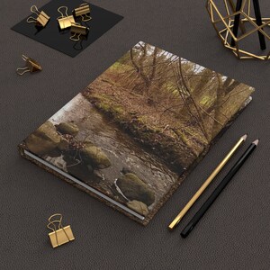 May include: A brown leather-bound journal with a nature scene on the cover. The scene features a stream flowing through a forest with rocks and trees.
