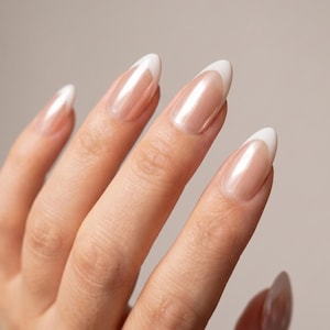 Pode incluir: Uma mão com unhas longas e ovais. Cada unha apresenta uma base perolada com uma ponta branca, criando um visual clássico de manicure francesa. As unhas são bem cuidadas e brilhantes.