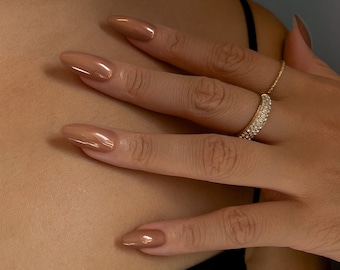 Könnte beinhalten: Nahaufnahme einer Hand mit langen, mandelförmigen, braunen Nägeln. Die Hand ist mit zwei Ringen geschmückt, einem mit einer Reihe kleiner, klarer Steine und einem weiteren schlichten Goldband. Die Person trägt ein schwarzes Oberteil.