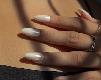 May include: Close-up of a hand with long, oval-shaped nails painted with a white and iridescent ombre nail polish.