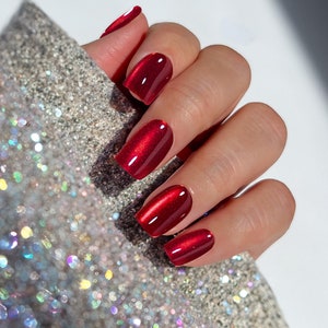 May include: Close-up of a hand with red nail polish. The nails are short and square-shaped, with a glossy finish. The hand is resting on a sparkly silver surface.
