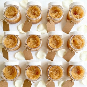 May include: An overhead shot shows twelve small, clear jars filled with a light brown, granular substance. Each jar has a white lid and a brown tag tied with twine. White petals are scattered around the jars on a white surface.