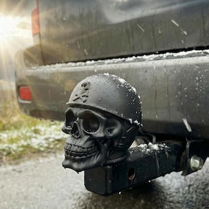 Op de afbeelding: Zwarte trekhaakafdekking in de vorm van een schedel met een militair helmdesign. De trekhaakafdekking is gemonteerd op een zwart voertuig, met de tekst "Wetterfest & UV-beständig - langlebiger Schutz" in het wit.