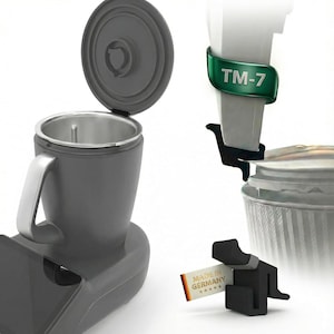 Könnte beinhalten: Eine dunkelgraue Kaffeemaschine mit Edelstahl-Innenraum und Griff. Der Deckel ist geöffnet und gibt den Blick ins Innere frei. Ein weißes und grünes Etikett trägt die Aufschrift "TM-7". Ein kleines Etikett trägt die Aufschrift "Made in Germany".